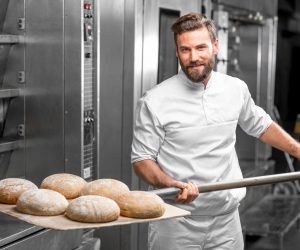 Handsome baker in uniform taking out with shovel freshly baked buckweat bread from the oven at the manufacturing